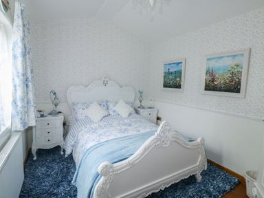 A bedroom with a white ornate bed blue floral bedding two nightstands and floral paintings at Turnpike Lodge in Brixham
