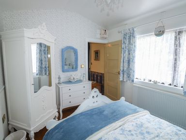 A bedroom with a white wardrobe a dresser and mirror a bed with blue bedding and floral curtains at Turnpike Lodge in Brixham