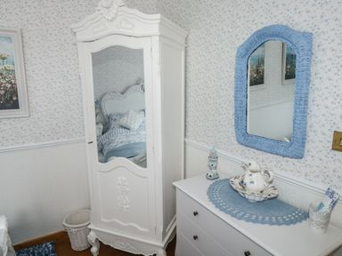 A bedroom corner with a white wardrobe with mirror a white dresser with a blue doily and framed mirror at Turnpike Lodge in Brixham