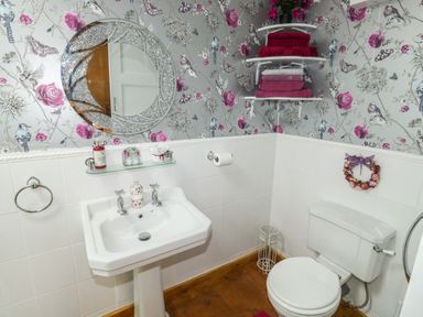 A bathroom with a pedestal sink a toilet a round mirror floral wallpaper and shelves with folded towels at Turnpike Lodge in Brixham