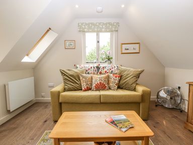 A small sitting area with a sofa adorned with cushions a wooden coffee table with magazines a window and a fan on the floor at The Attic at Redmond Bottom in Wookey