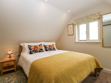 A bedroom with a bed covered in a yellow quilt two floral pillows a wooden nightstand and a window with a patterned blind at The Attic at Redmond Bottom in Wookey