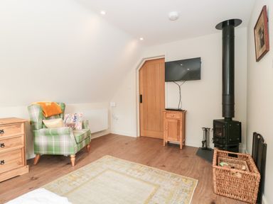A room with a plaid armchair next to a wooden dresser a small wooden cabinet under a wall-mounted TV a wood-burning stove and a wicker basket with firewood at The Attic at Redmond Bottom in Wookey