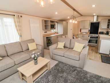 A living room with beige sofas and yellow pillows a wooden coffee table with flowers and an open kitchen with dining area at 20 Wolds View East Heslerton near West Heslerton
