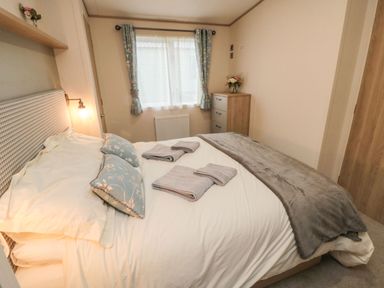 A bedroom with a bed linens and pillows with blue floral cushions towels on bed a window with blue curtains and a chest of drawers at 20 Wolds View East Heslerton near West Heslerton