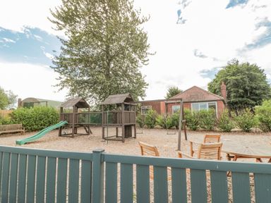 A fenced playground with wooden play structures swings slide and outdoor furniture near a brick building at 20 Wolds View East Heslerton near West Heslerton