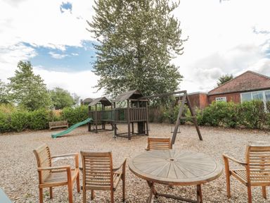 A playground area with wooden play structures and a slide surrounded by wooden chairs and a round table at 20 Wolds View in East Heslerton near West Heslerton