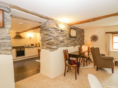 A living area with wooden chairs a small table and a plaid armchair next to a window and an open kitchen with appliances at Myra's View in Newby Bridge