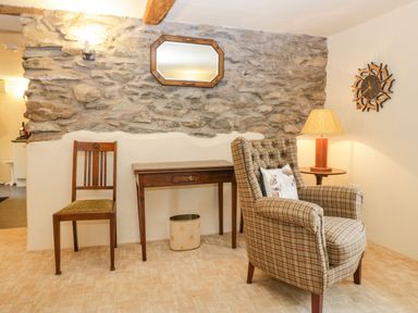 A room with a wooden chair, a small wooden table, a plaid armchair with a pillow, a lamp on a side table, a wall clock, and a stone wall with a mirror at Myra's View in Newby Bridge