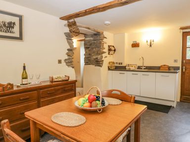 A kitchen with a wooden dining table and chairs a fruit basket on the table a wooden sideboard with wine and glasses white cabinets with a sink a wooden door exposed stone wall and ceiling beams at Myra's View in Newby Bridge