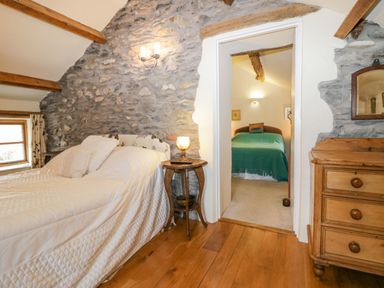 A bedroom with a bed covered with white bedding and a wooden nightstand next to a stone wall and a doorway leading to another room with a bed covered with a green blanket at Myra's View in Newby Bridge