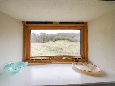 A small window with a wooden frame showing an outdoor grassy field with trees and a shelf under the window holding a blue glass decorative bowl and a floral plate at Myra's View in Newby Bridge
