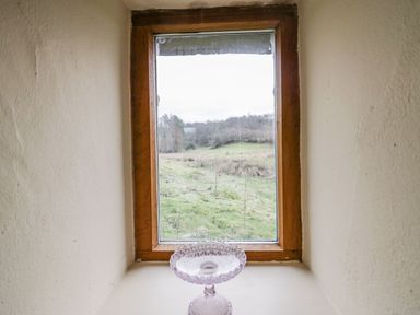 A small window with a wooden frame overlooking a grassy field with trees beyond and a glass pedestal bowl on the windowsill at Myra's View in Newby Bridge