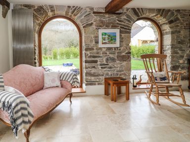 A sitting area with a pink sofa and wooden rocking chair near arched stone windows at Birchenhayes in St Dominick