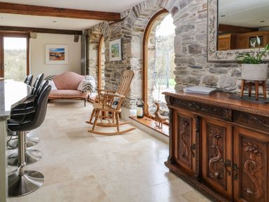 A stone wall room with arched windows bar stools rocking chair pink loveseat and wooden sideboard at Birchenhayes in St Dominick