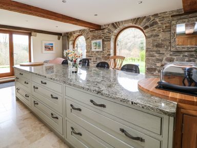A kitchen island with granite countertop and chairs in a room with stone walls and arched windows at Birchenhayes in St Dominick