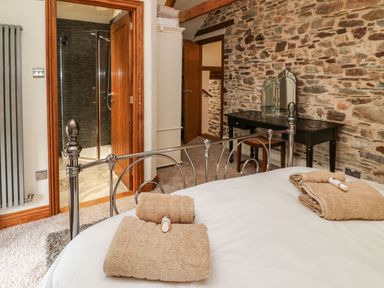 A bedroom with a metal bed frame folded towels with toiletries on the bed a stone wall a black dressing table with a mirror a wooden door leading to a bathroom with a shower at Birchenhayes in St Dominick