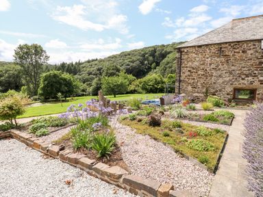 A stone garden with flower beds next to a stone building and a green lawn with trees at Birchenhayes in St Dominick