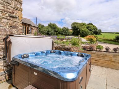 An outdoor hot tub next to a stone building with a garden and trees in the background at Birchenhayes in St Dominick