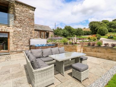 An outdoor patio with wicker furniture and cushions next to a stone building and garden at Birchenhayes St Dominick