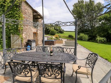 A patio with a metal table and chairs set under a pergola near a stone house with a barbecue grill and a hot tub in a garden at Birchenhayes in St Dominick