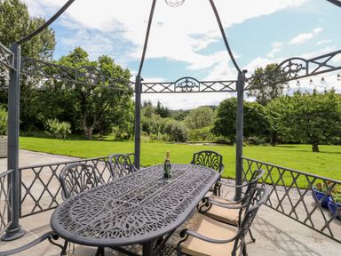 An outdoor metal table and chairs under a gazebo with a bottle on the table and a green garden in the background at Birchenhayes in St Dominick