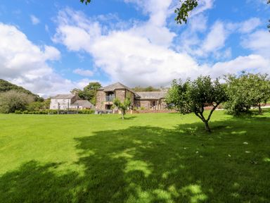 A grassy garden with small trees and stone houses under a partly cloudy sky at Birchenhayes in St Dominick