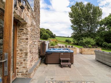 An outdoor patio area with a hot tub wooden steps stone wall doorway and a sofa beside wooden planters and trees at Birchenhayes in St Dominick