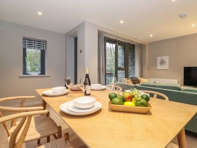 A dining table with plates glasses a bottle and fruit bowl next to a living area with sofas and large windows at Bruach in Backbarrow