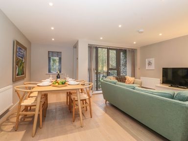 A dining table with six chairs set with bowls and glasses next to a living area with a green sofa and a television at Bruach in Backbarrow