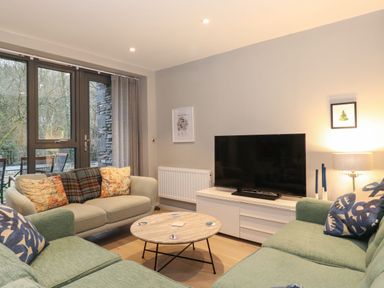 A living room with green and beige sofas around a round wooden coffee table and a TV on a white stand at Bruach in Backbarrow