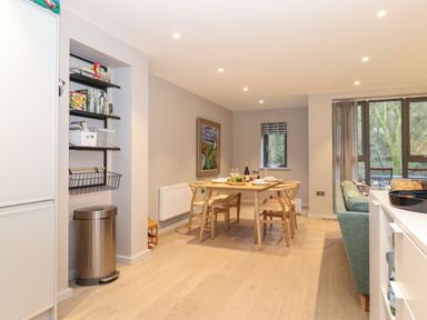 A dining area with a wooden table set with dishes and wine glasses next to a living room with a green sofa and large windows at Bruach in Backbarrow