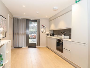 A kitchen with white cabinets wooden floor and a glass door with grey curtains at Bruach in Backbarrow