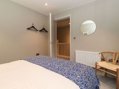 A bedroom with a bed covered in a patterned blue and white blanket a wooden chair round mirror on the wall and two black hangers on the opposite wall at Bruach in Backbarrow