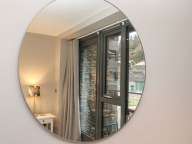A round mirror reflecting a window with stone wall outside a curtain and a table lamp on a side table at Bruach in Backbarrow