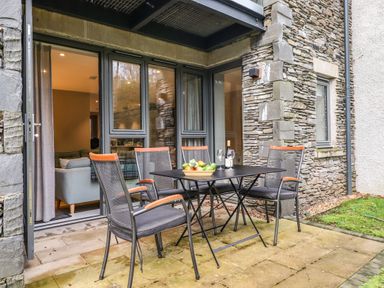 An outdoor patio area with a black table and four chairs next to a stone wall and glass doors at Bruach in Backbarrow