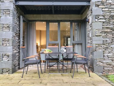 An outdoor patio with a table and four chairs in front of stone walls and glass doors at Bruach in Backbarrow