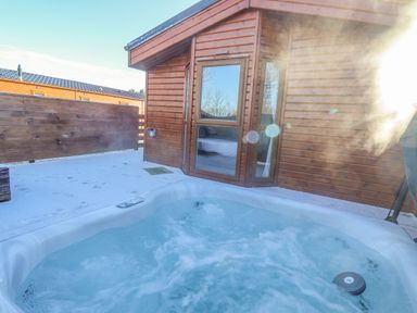 Outdoor hot tub on a snowy patio next to a wooden cabin with a glass door at Pegasus in Felmoor Park