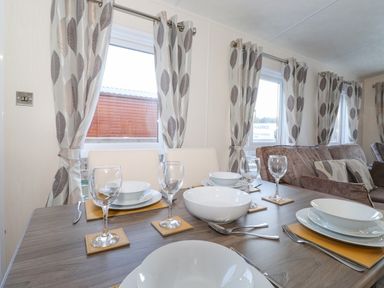 A dining table set with plates bowls glasses and cutlery next to windows with patterned curtains and a sofa in the background at Pegasus in Felmoor Park