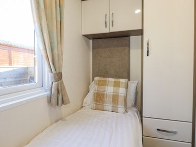 A single bed with a checked cushion near a window with curtains and white cabinets in a bedroom at Pegasus in Felmoor Park