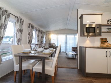 A dining area with a table set for six next to windows with patterned curtains and a kitchen with a microwave at Hercules Felmoor Park