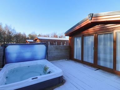 An outdoor deck area with a hot tub and wooden cabins in the background at Canis in Felmoor Park