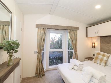 A bedroom with a bed with folded towels and a checkered pillow a bay window with curtains and a vase with green leaves on a shelf at Canis in Felmoor Park