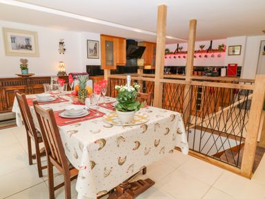 A dining area with a table set for six with a tablecloth featuring chickens and a kitchen with wooden cabinets in the background at Monkleigh Mill in Monkleigh near Bideford