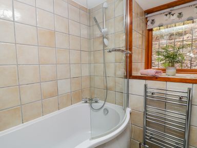 A bathroom with a white bathtub tiled walls a glass shower screen a window with a plant and towel and a chrome heated towel rail at Monkleigh Mill Monkleigh near Bideford