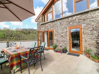 An outdoor patio with a table covered by a checkered tablecloth set with plates and glasses next to a stone building with wooden framed glass doors and potted plants at Monkleigh Mill in Monkleigh near Bideford