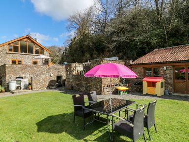 An outdoor garden area with a glass table and four chairs under a pink umbrella next to stone buildings with plants and a children's playhouse at Monkleigh Mill in Monkleigh near Bideford