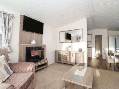A living room with a sofa, coffee table, mounted TV, cabinet with mirror, and an open view into a dining area at Draco Felmoor Park