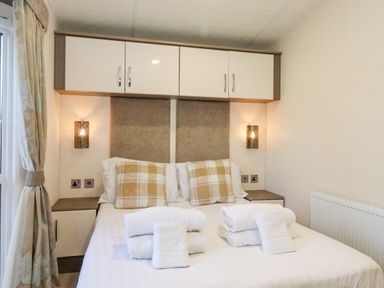 A bedroom with a double bed pillows and folded towels bedside tables wall lamps overhead cabinets and curtains at Draco in Felmoor Park