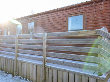 A wooden cabin with windows and a door behind a wooden railing with snow on the ground and railing at Draco in Felmoor Park